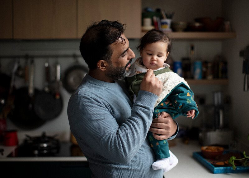 Hatem, an Iraqi refugee, is seen in a kitchen in South London, UK, holding Khairun's baby daughter. Hatem smiles warmly at the curious baby, whose face is illuminated by a lovely light streaming in from a front window