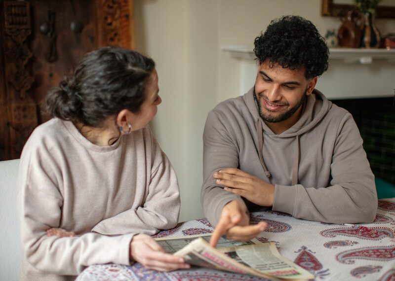 Two individuals, Belén and Anes, are seated indoors at a table, happily reviewing newspaper articles about Anes's family's resettlement in South London, UK