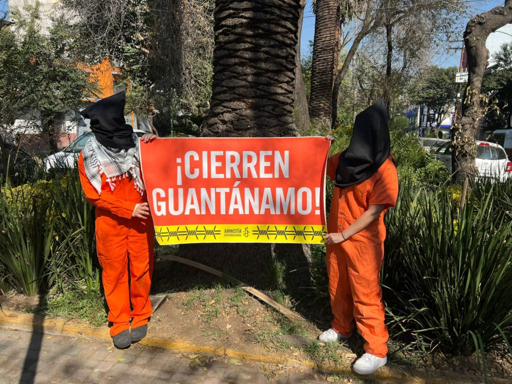 ناشطان يرتديان بذلات برتقالية اللون وأكياسًا سوداء على رأسيهما، يحملان لافتة مكتوب عليها