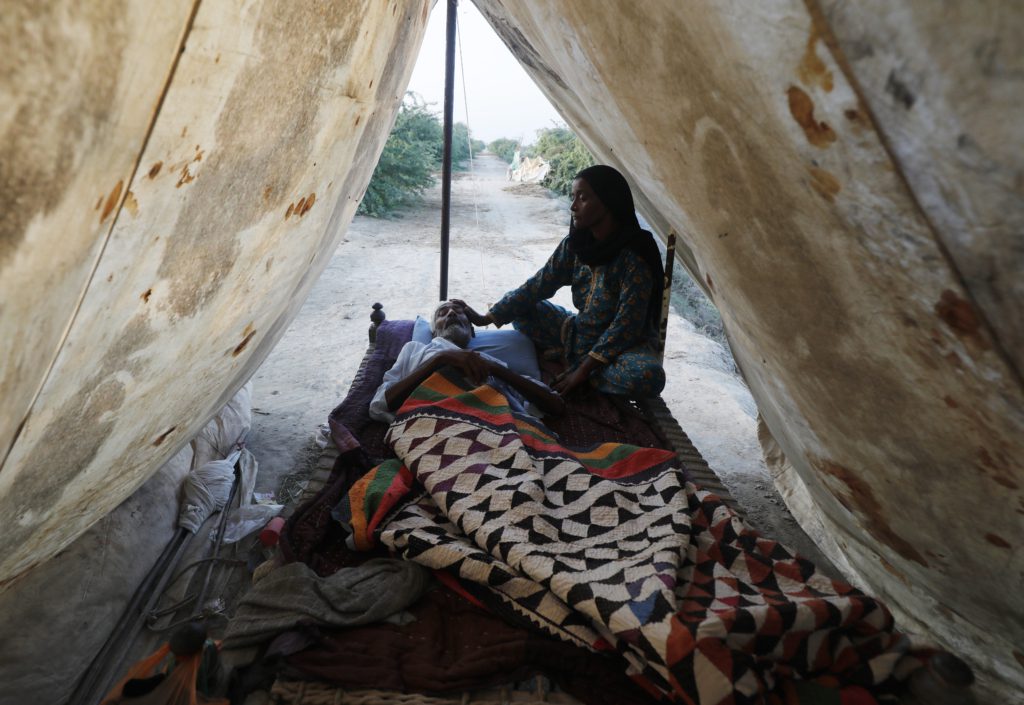 A photo of an ill elderly man, Haji, lying down in a tent accompanied by his wife who is sitting next to him with her hand on his temple.