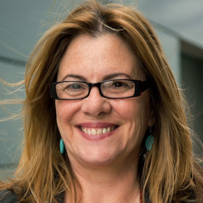 Ann is smiling. She is wearing black framed glasses and turquoise earrings.