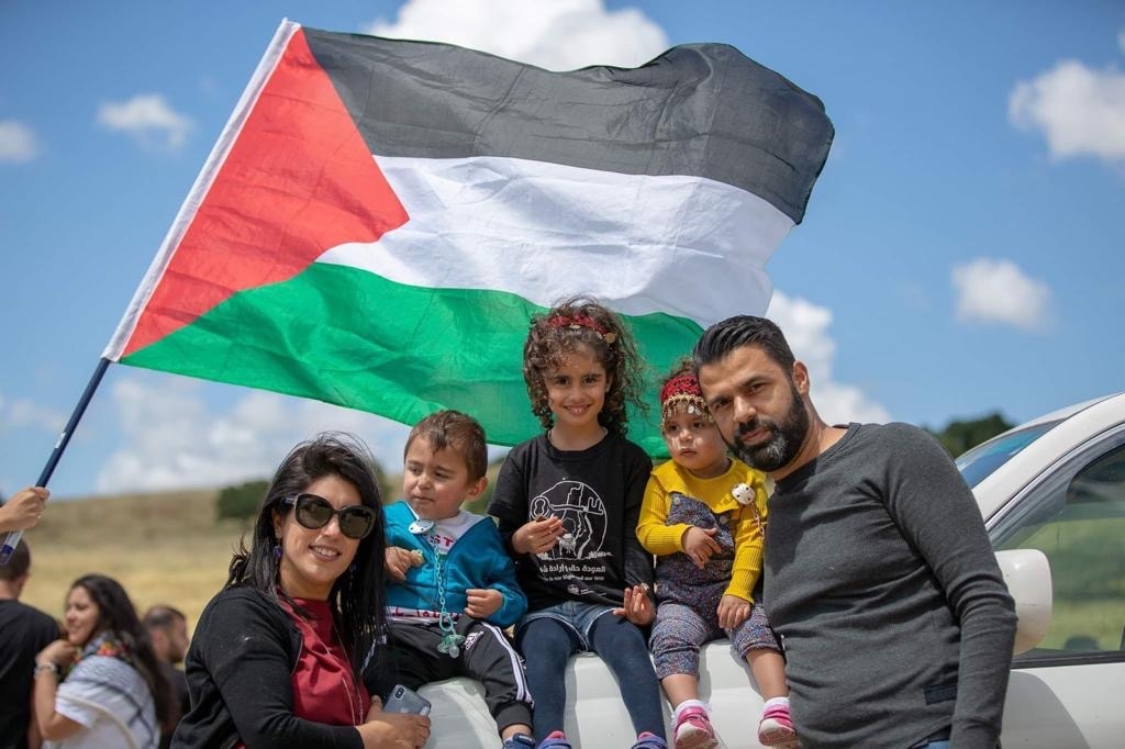 A family of four sit on a white car, with a Palestine flag blowing in the background.