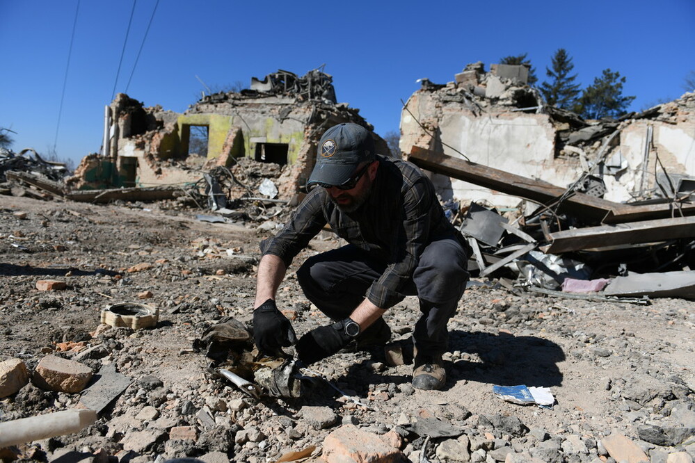 Brian, wearing black clothes, sunglasses, a hat and gloves. He is picking up remnants of a weapon found amid the rubble in Ukraine 