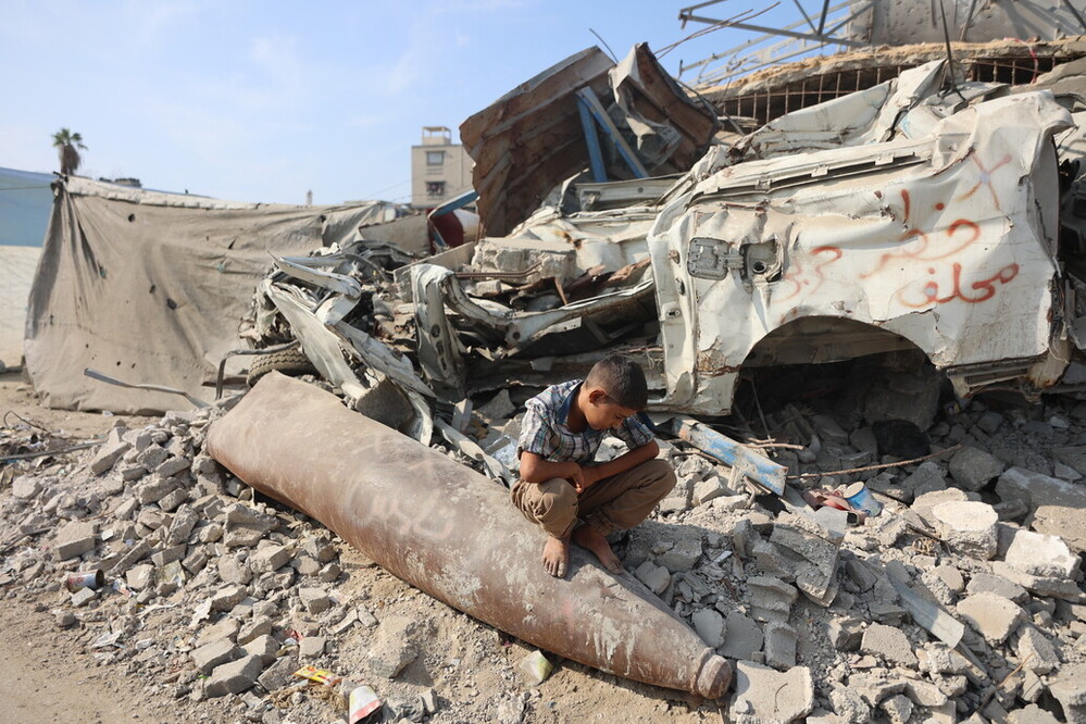 a young boy sits on an unexploded missile in Gaza. He is surrounded by rubble from destroyed buildings.