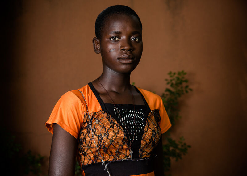 A young girl, wearing an orange dress, stands looking to the camera.