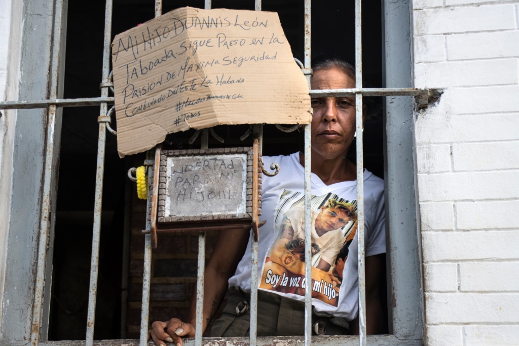 Yenisey standing behind bars with a cardboard placard. She is wearing a white t shirt with a picture of her son Luz. 