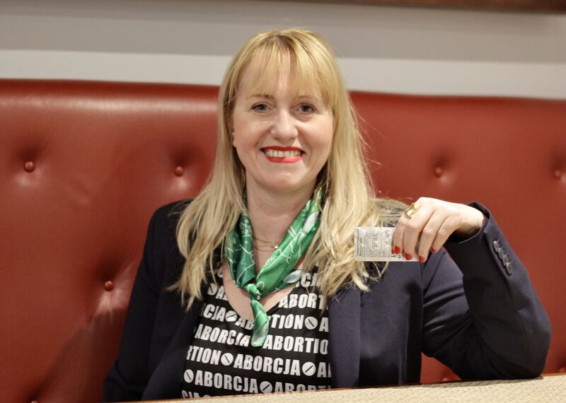 Kinga Jelińska, activist from Poland looks at the camera as she is holding a blister pack of medication. She has blond hair and is wearing a black jacket and a green scarf.