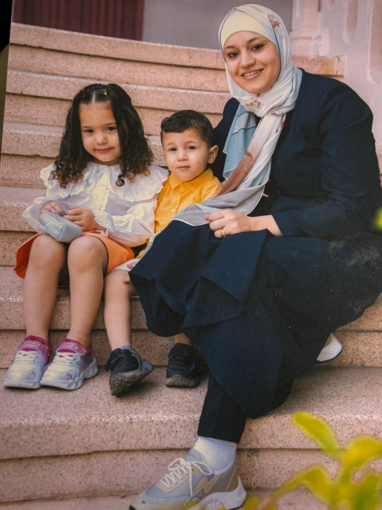 Una madre, sentada en unos escalones color melocot&oacute;n moteados, junto a su hija e hijo peque&ntilde;os. Sonr&iacute;en a la c&aacute;mara.