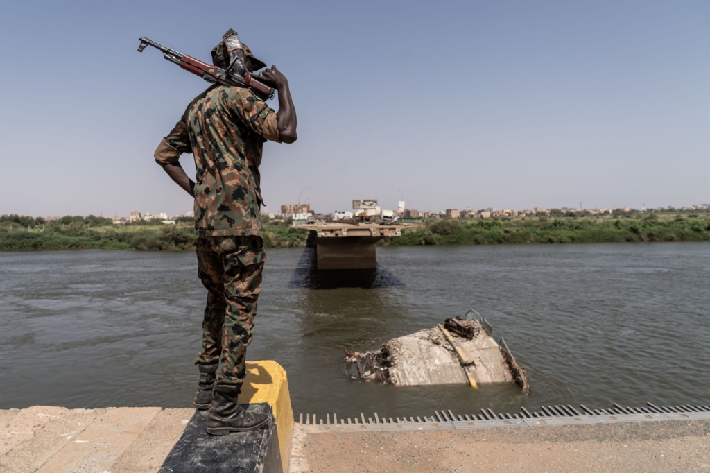 a soldier holding a gun over his shoulder, standing at the end of a road where a bridge has been destroyed. You can see a part of the bridge submerged underwater.