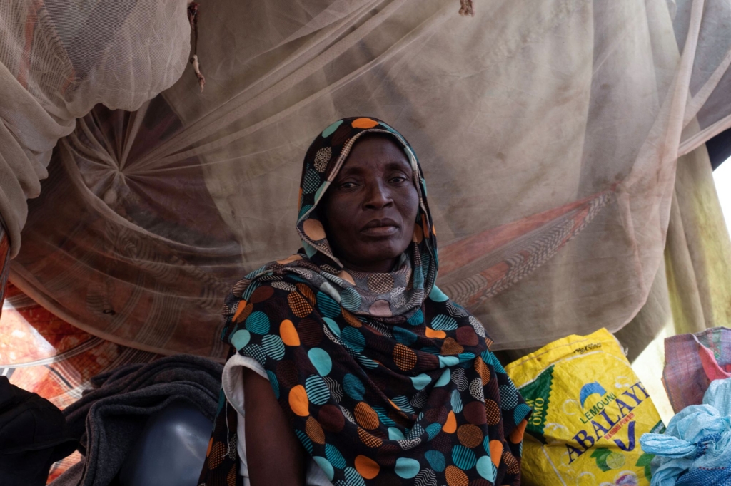 a portrait of a woman at a camp for displaced people. she has a solemn expression as she looks to the camera. behind her, there is fabric draped from the sides of her temporary home. 