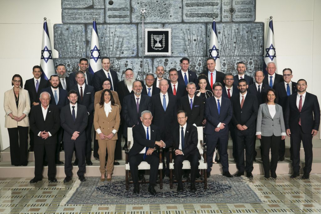 Members of cabinet stand in front of flags of Israel, while two men in the front of them shake hands