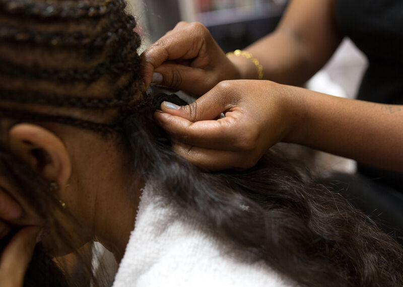 A woman leans forward as she has her hair braided by a person's hands