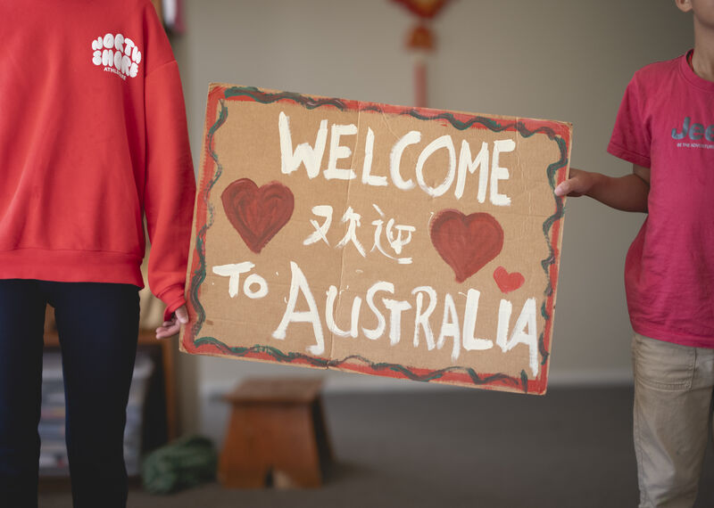 Two children stand between a hand painted sign reading "welcome to Australia"