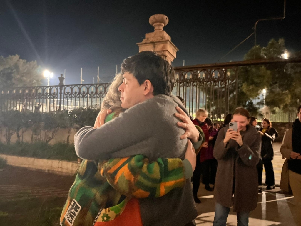 A man in a grey jumper warmly embraces an older woman outside an official looking building.