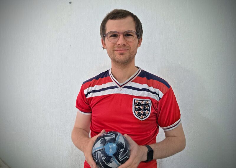 A gentleman, wearing glasses and a red England football shirt, smiles to camera, as he holds a football