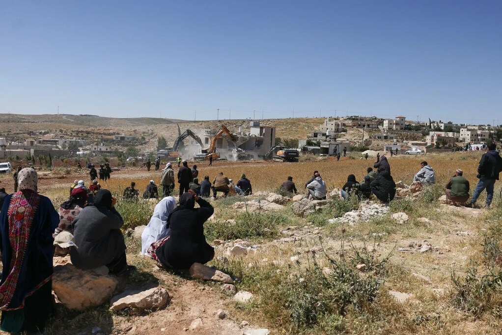 People sitting on rocks look towards two excavators demolishing a structure