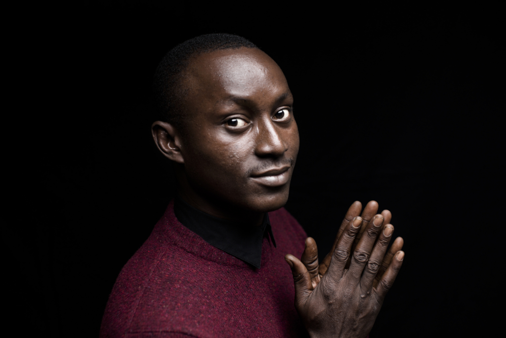 A man stands looking to camera with his hands clasped. He wears a red jumper.
