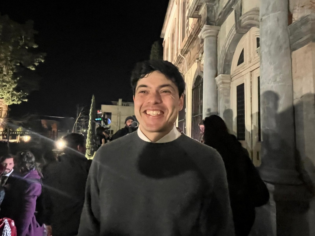 As night closes in, a man in a dark jumper stands against a brightly lit building.