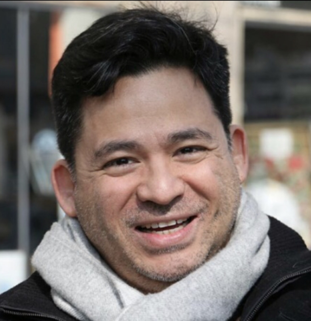 Jose's headshot looking at the camara with a smile, light grey headscarf