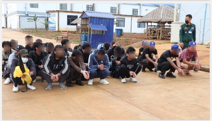A group of people in a scamming compound sit on the floor during a police raid, some of them are Leap Somnang security guards in blue hats.