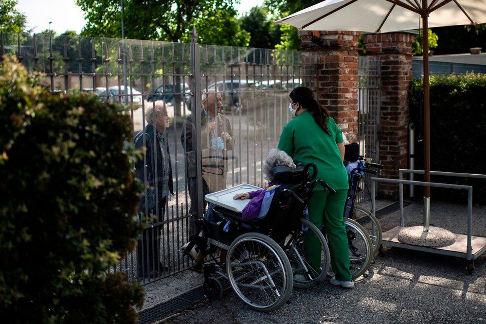 Una mujer en silla de ruedas y una trabajadora de una residencia de personas mayores hablan desde dentro con las personas que se encuentran al otro lado de la puerta de entrada. Una barrera protectora cubre la verja para evitar la propagación del virus.