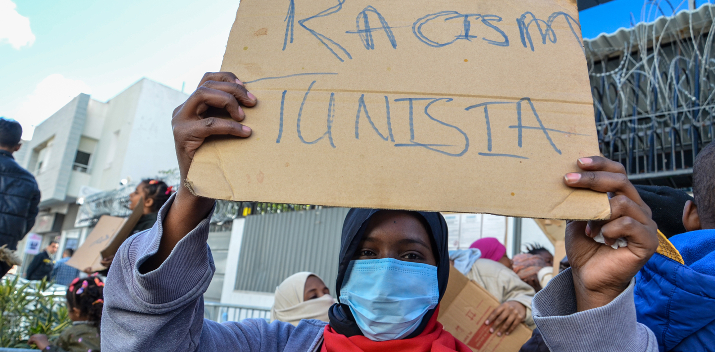 Persona negra con mascarilla sostiene un cartel sobre la cabeza que dice “Racismo Túnez”.