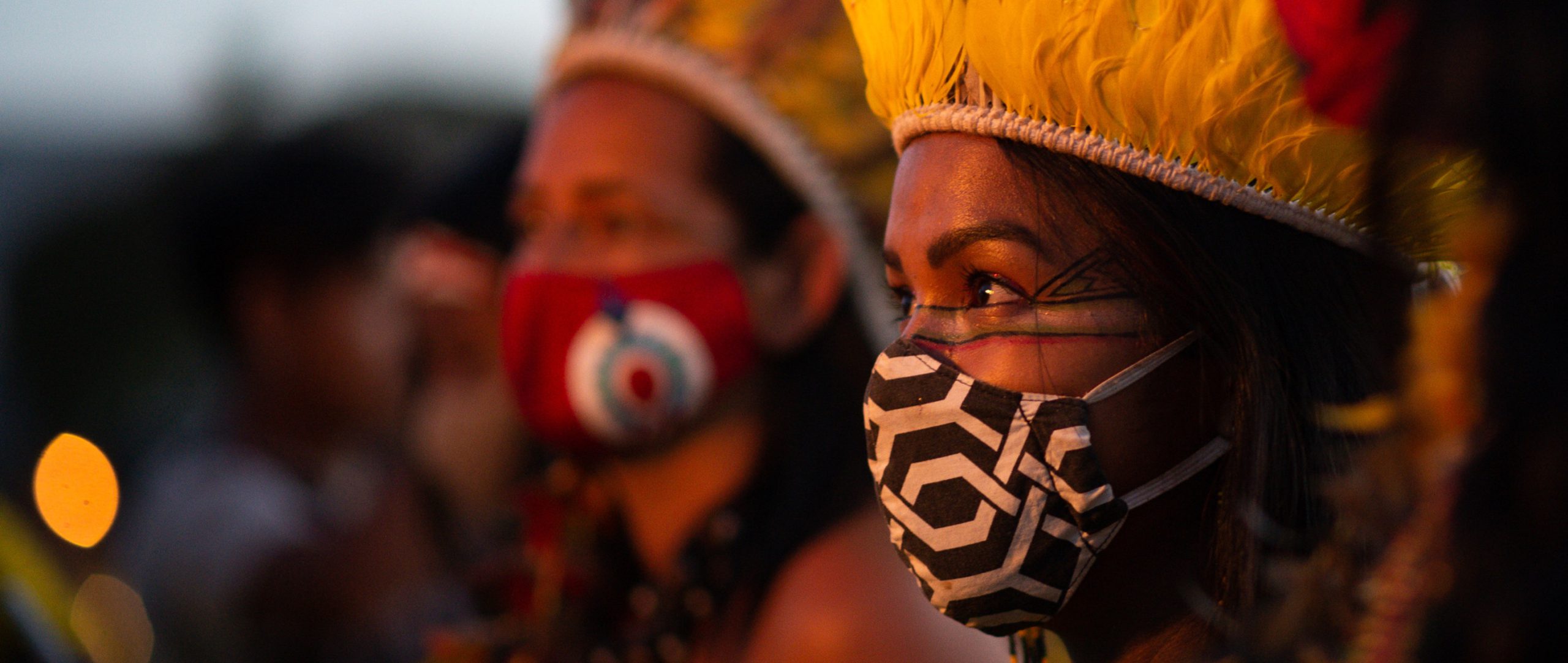 Personas en fila con mascarillas estampadas y vistosos tocados de plumas.