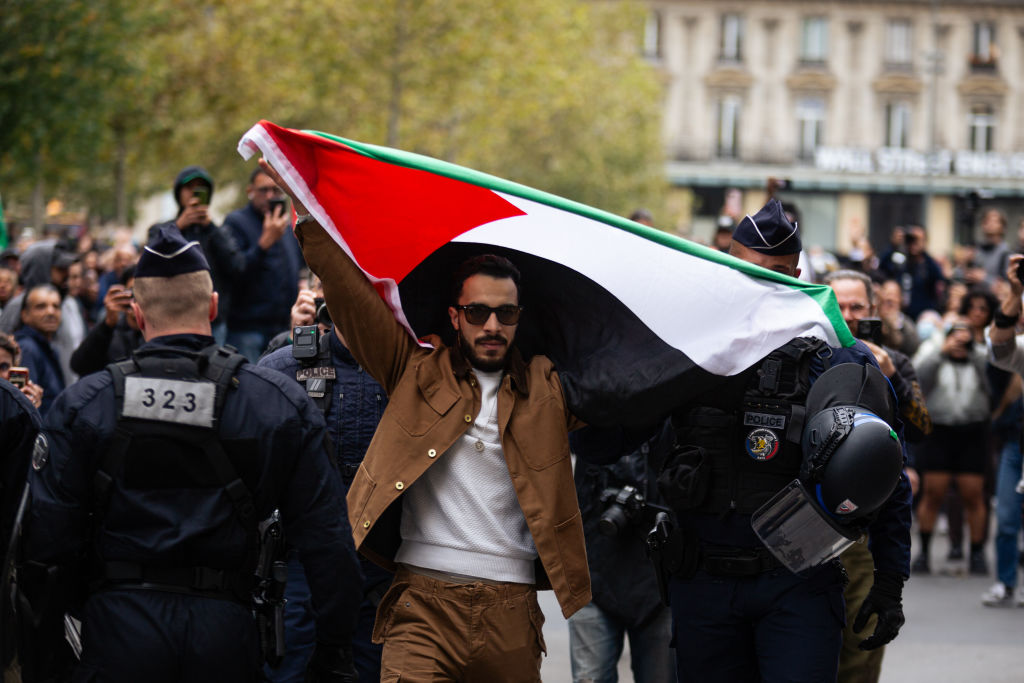 Un hombre vestido con chaqueta y pantalón de color tostado sostiene en alto una bandera palestina. Está rodeado de policías.