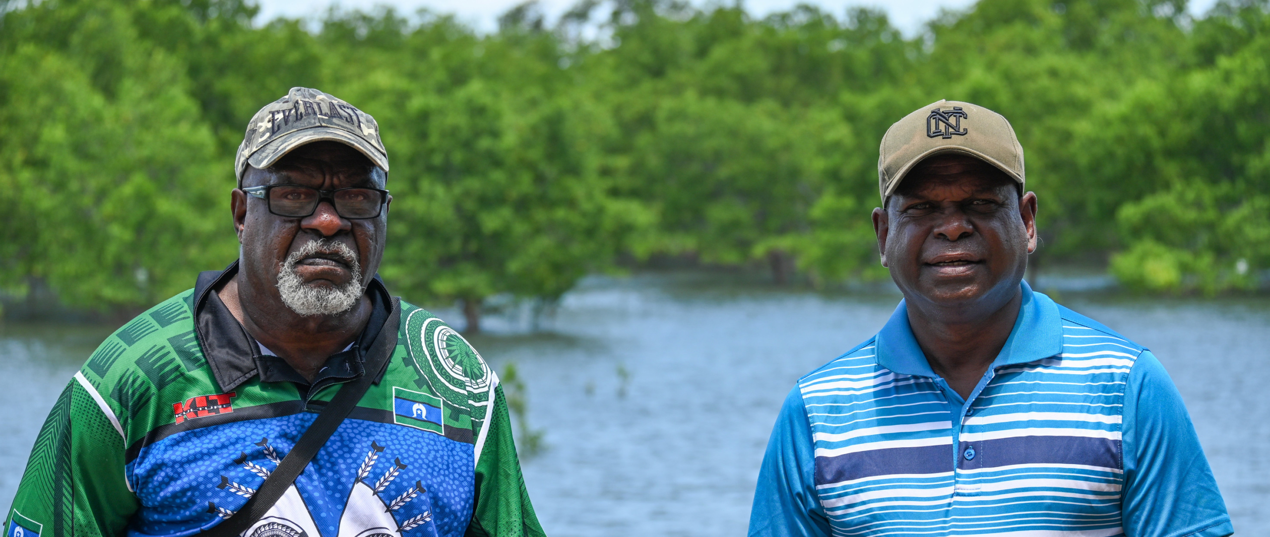 Uncle Pabai y Uncle Paul, líderes comunitarios de la nación Guda Maluyligal, posan junto a un río y frondosa vegetación.