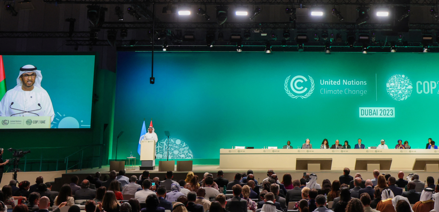 Imagen del interior del salón de conferencias de la COP28 con el presidente de la cumbre, Sulan Al Jaber, dirigiéndose a los asistentes sobre un fondo verde y el logotipo de la COP28.
