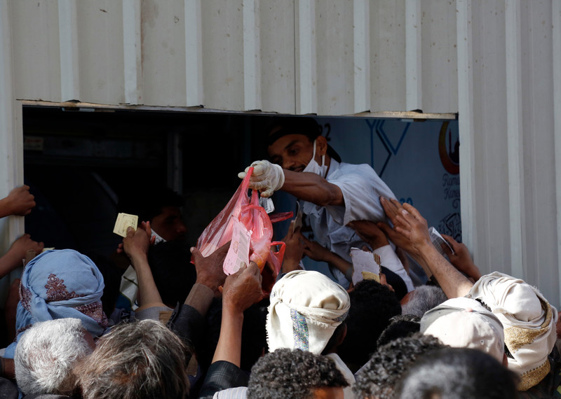 Una multitud extiende las manos hacia una bolsa de plástico con comida que ofrece un trabajador uniformado detrás de una ventanilla.