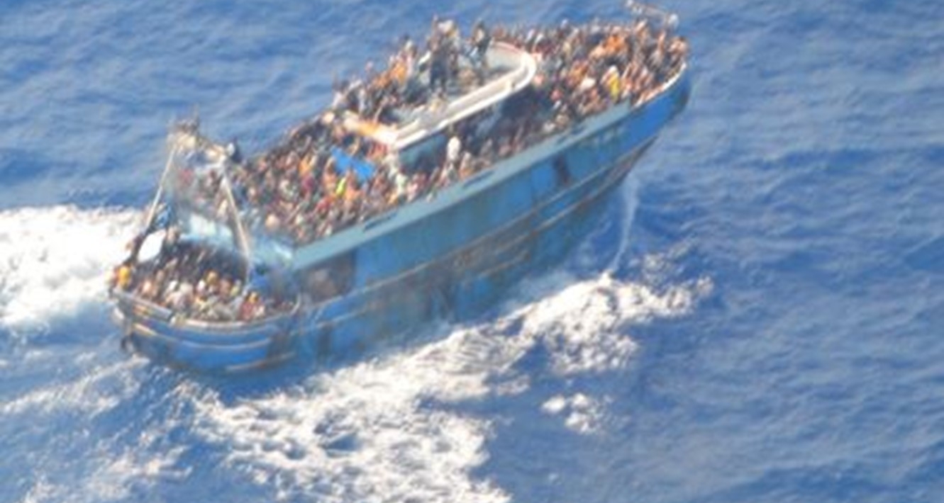 e ve un barco azul abarrotado de gente sobre un mar azul. La imagen es una vista aérea. La espuma blanca indica que el barco se mueve. Se ve gente en todas las cubiertas.