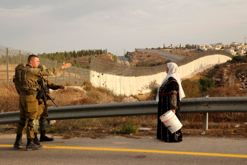Una mujer palestina espera de pie, junto a otras personas (a las que no se ve en la foto), cerca de un puesto de control del ejército israelí, a poder pasar a sus campos de olivos situados al otro lado de la barrera de separación de Israel (fondo de la imagen) tras haber recibido un permiso especial para recolectar la aceituna, el 13 de octubre de 2021, cerca de la población de Bait Awa, a las afueras de la ciudad cisjordana de Hebrón.