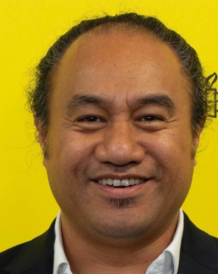 Peter Fa'afui sobre un fondo amarillo con el logo de Amnistía. Viste camisa blanca y chaqueta. 