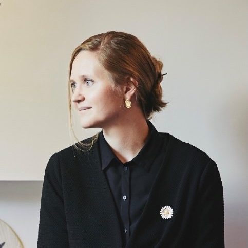 Nina Boel sentada con una pared blanca de fondo y la cabeza girada hacia la izquierda. Viste camisa negra y chaqueta de punto. 