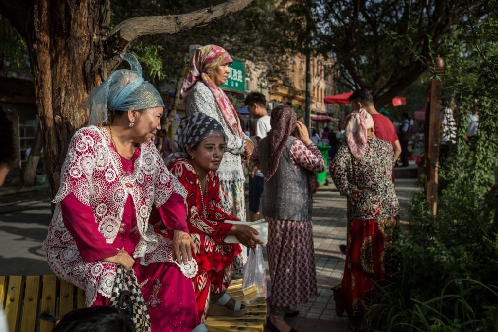 Dos mujeres están sentadas en un banco. Ambas visten ropas y pañuelos profusamente adornados.