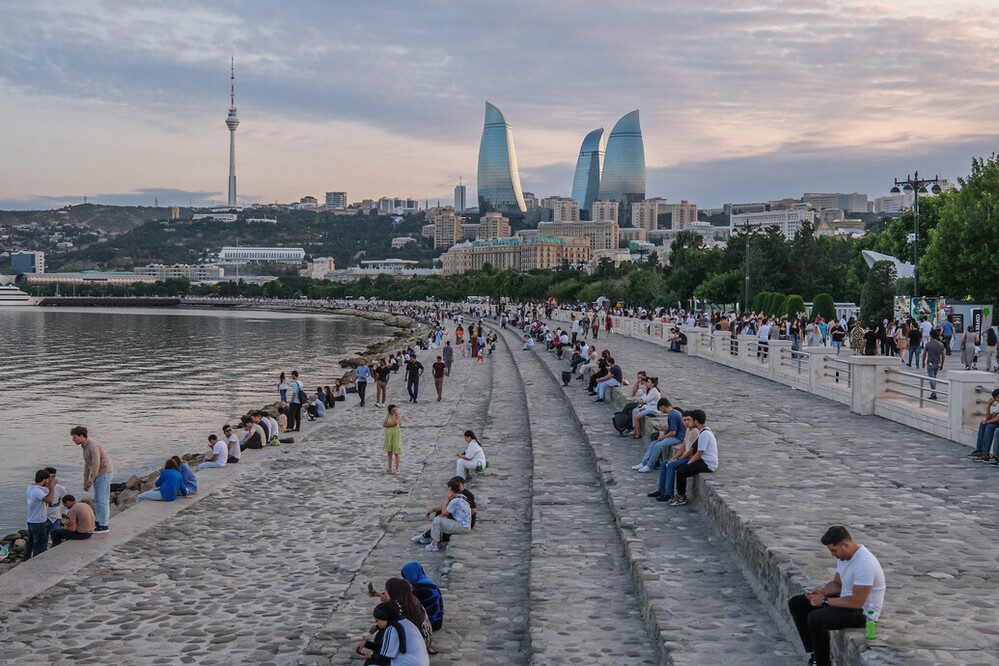 a collection of people sit on stone steps with a few of the Caspian sea. The Baka skyline can be seen on the horizon.