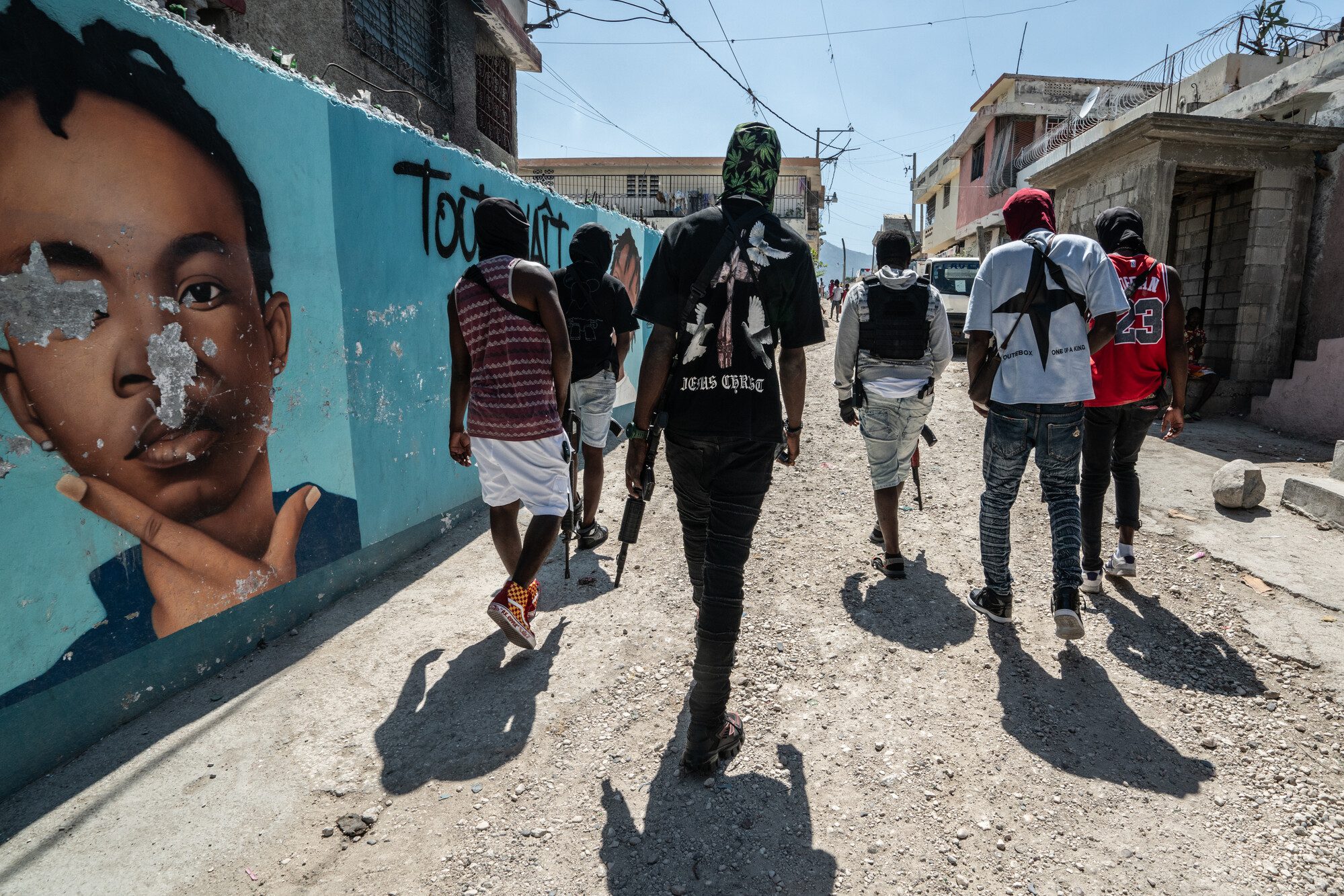 Un grupo de miembros armados de una banda criminal caminan juntos. Llevan las armas colgadas a la espalda y algunos llevan chalecos antibalas. Todos van enmascarados. Pasan junto a un mural con la imagen de un niño.