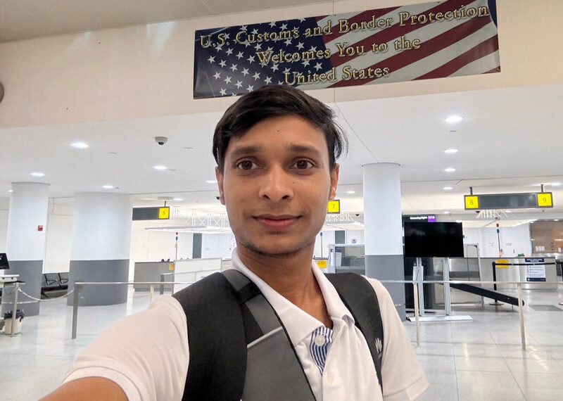 Sawyeddollah en un aeropuerto delante de la bandera estadounidense. En la bandera se lee: “La Oficina de Aduanas y Protección Fronteriza le da la bienvenida a los Estados Unidos”.