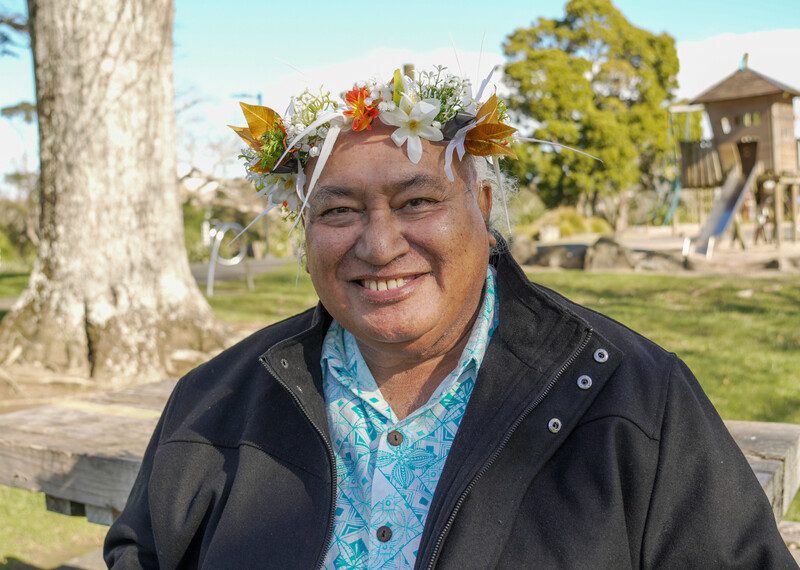 Un hombre con una corona de flores sonríe sentado en un banco al aire libre.