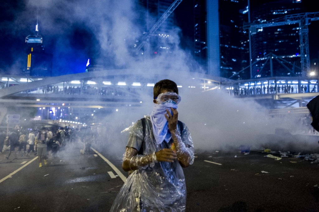 Un manifestante, con elementos de protección como gafas y un paño cubriéndole la cara, camina por una calle; detrás puede verse una nube de gas lacrimógeno.
