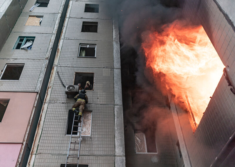 Un bombero asciende por una escalera hacia dos personas asomadas a una de las ventanas de un edificio alto envuelto en un gran incendio.