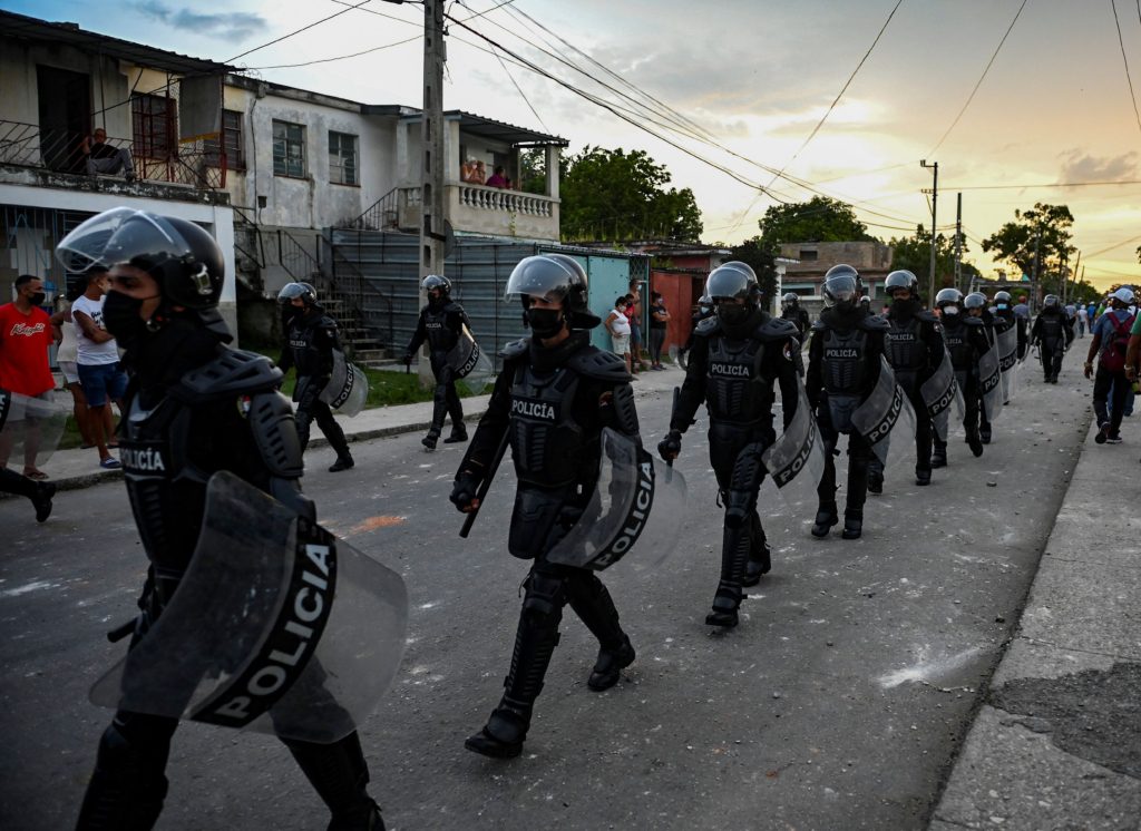 Policías antidisturbios fuertemente armados caminan en fila india por una calle. Todos ellos llevan cascos con visera y grandes escudos antidisturbios en el brazo izquierdo.