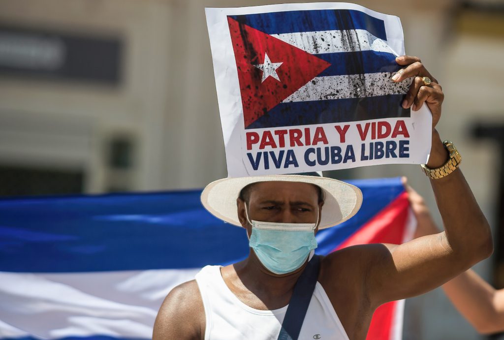Manifestante con una mascarilla sujetando un letrero que dice “Patria y Vida, Viva Cuba Libre” junto con la bandera cubana.