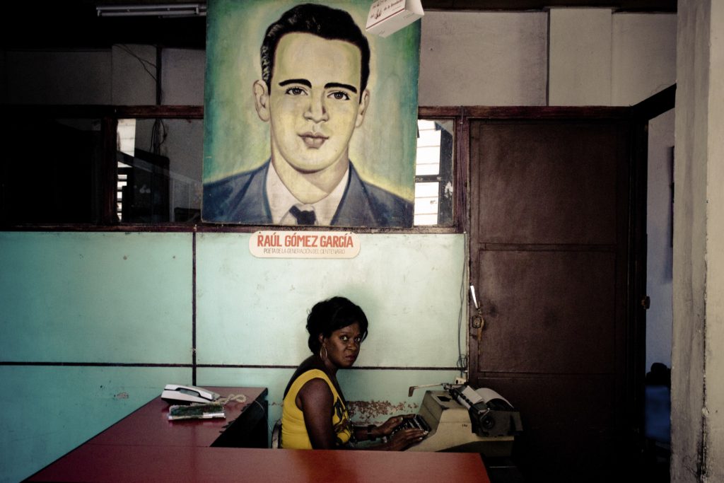 Una oficinista cubana mecanografía en un escritorio bajo un gran retrato colorido del líder revolucionario Raúl Gómez García colgado sobre una pared turquesa.