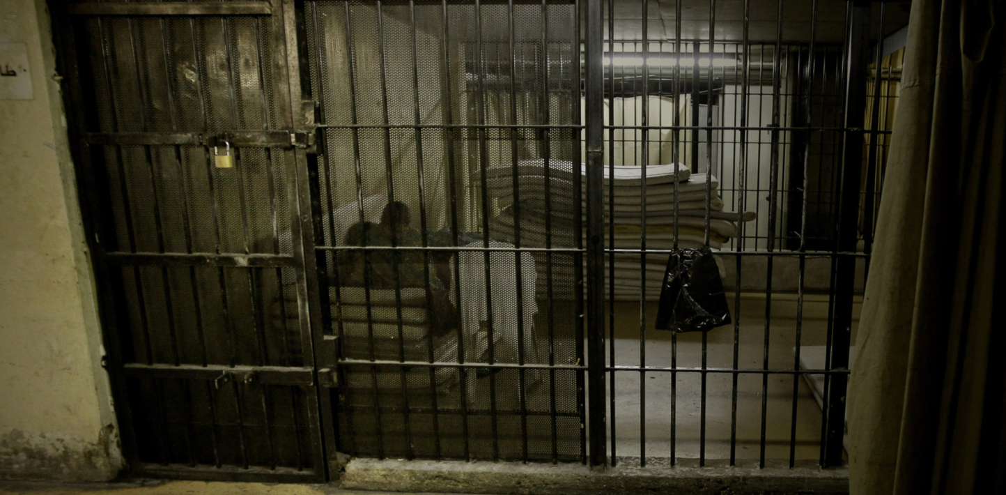 A man sits in a dingy cell, behind prison bars with flourescent lighting on the back wall.