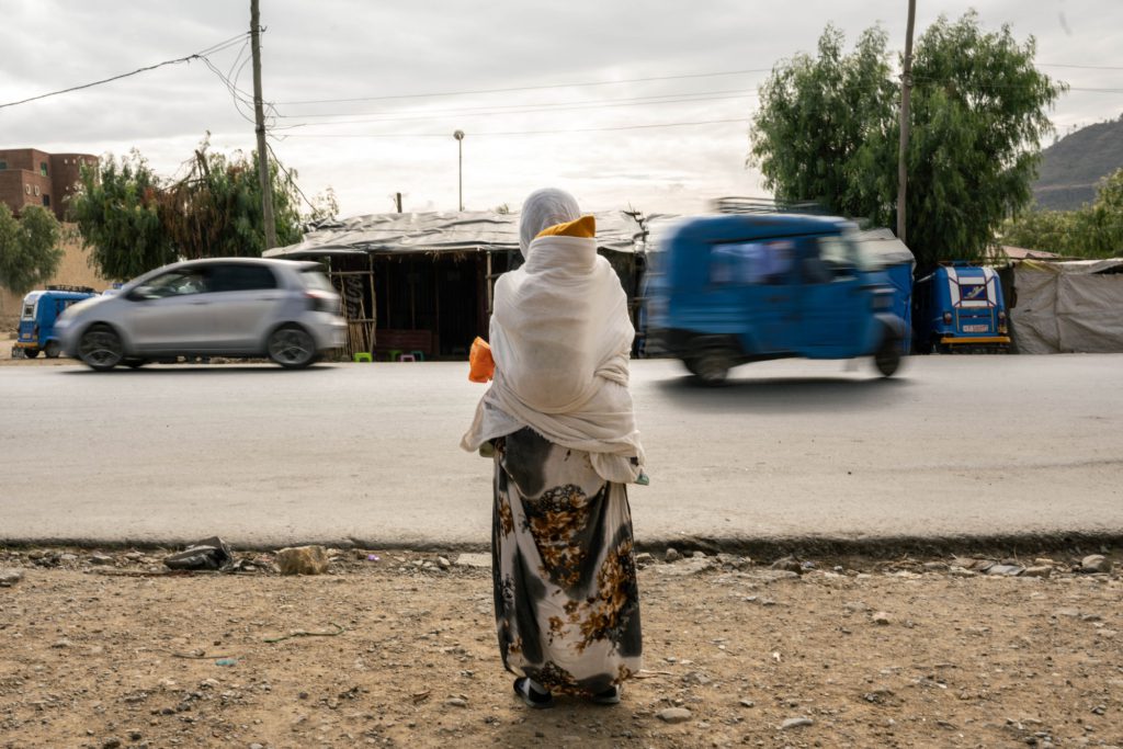 A woman stands on the side of the road as cars pass by. A small child is strapped to her back.