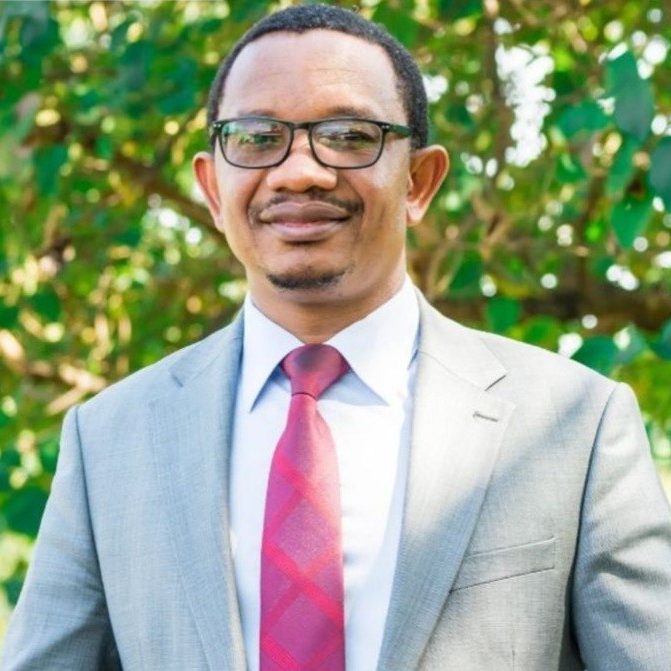Godfrey is standing in front of green foliage wearing a grey suit and glasses.