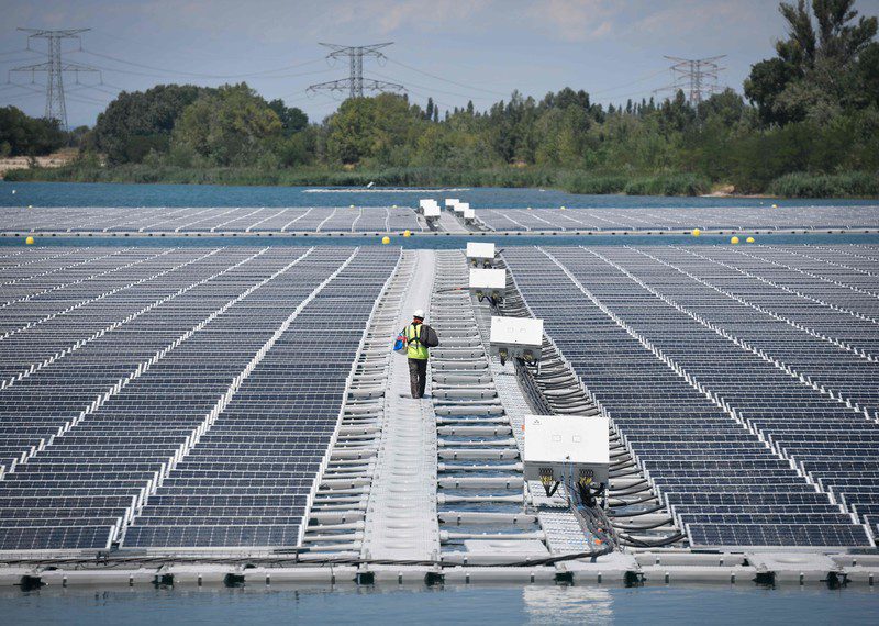 a worker in a white helmet and a high visibility jacket walks amid rows of solar panels.