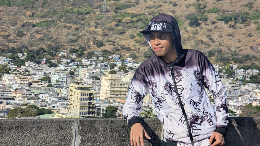A young man in a white and black hooded top, stands against a city backdrop, smiling. He looks cool, as young people do.